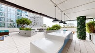 Located on the 4th floor of 7 Lime Ave in Long Beach, California, this expansive rooftop terrace offers a well-appointed outdoor amenity space with a modern, resort-inspired design. The terrace is organized into several distinct zones, each thoughtfully designed for socializing and relaxation. At its centerpiece sits a circular fire pit surrounded by a generous wraparound bench with blue cushioned seating and matching bar-height chairs, creating an inviting gathering focal point. A covered bar area features a sleek white countertop with blue panel detailing, bar stools, pendant lighting, a wall-mounted television, and vertical green wall accents, offering an ideal space for casual entertaining. A separate lounge nook provides teal-cushioned outdoor seating framed by lush mature trees and planters. Large concrete raised planters throughout the terrace are filled with vibrant greenery, adding a natural and organic contrast to the clean, modern architecture. An extended walkway along the perimeter of the terrace provides panoramic views of the Long Beach skyline, palm tree-lined streets, and neighboring high-rises. The consistent use of light-toned large-format tile flooring, white composite decking, and crisp white walls ties the entire terrace together, projecting a bright, airy, and contemporary aesthetic throughout.