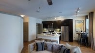 This single-floor residence showcases an open-concept layout that integrates the kitchen and living area into one cohesive, functional space. The kitchen is a standout feature, outfitted with rich, dark espresso cabinetry that extends to the ceiling, providing generous storage throughout. Stainless steel appliances, including a full-size refrigerator, microwave, and range, lend a sleek, modern aesthetic. The kitchen island serves as a natural focal point, topped with a light-toned marble-style surface and lined with bar-height stools, making it ideal for casual dining or entertaining. Track lighting overhead ensures the kitchen is well-illuminated, while a ceiling fan in the living area promotes air circulation. The living space is anchored by a tufted, neutral-toned sofa dressed with coordinating decorative pillows, creating a relaxed and inviting atmosphere. Hardwood-style flooring flows throughout, tying the areas together cohesively. A hallway visible in the background suggests access to additional private rooms, including what appears to be a closet or utility area. The overall ambiance is contemporary and well-appointed.