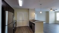 This single-floor space showcases a well-appointed, open-concept layout where the kitchen serves as a central hub. The kitchen is outfitted with dark cabinetry that contrasts elegantly against granite countertops, which extend across both the perimeter workspace and a peninsula island. The island features a stainless steel undermount sink and provides additional seating or prep space, making it both functional and social. Two pendant lights hang above the peninsula, adding focused ambient lighting that complements the recessed ceiling fixture overhead. Stainless steel appliances, including a refrigerator and dishwasher, complete the kitchen's modern aesthetic. A set of louvered double doors offers access to a closet or utility area, adding practical storage to the space. The warm-toned, hardwood-style flooring runs throughout, lending a cohesive and grounded feel to the interior. Soft yellow-green wall tones create a welcoming atmosphere, while the open sightlines from the kitchen into the adjoining living area enhance the sense of spaciousness. Natural and artificial lighting work together to keep the space bright and inviting throughout.
