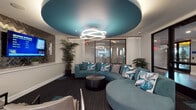 This single-floor space is thoughtfully designed with a contemporary aesthetic, combining functional areas with striking interior details. The lounge area serves as a central gathering point, distinguished by a bold teal circular ceiling feature with integrated ambient lighting and a sculptural multi-ring pendant light. Teal sectional and accent sofas are arranged around a round coffee table, while a wall-mounted display and a textured herringbone accent wall add visual depth. Tall indoor palms and sheer white curtains contribute to a polished, welcoming atmosphere. Glass-paneled partitions provide transparency between spaces without sacrificing definition.

The kitchen area is finished with large-format porcelain tile flooring, dark espresso cabinetry, and a generous island topped with veined white stone countertops. A textured white tile backsplash reflects light from the crystal chandelier overhead, while stainless steel appliances complete the functional layout. Pops of yellow on an adjacent accent wall introduce energy to the space.

The conference room accommodates a sizable dark wood table surrounded by upholstered yellow and charcoal task chairs. A wall-mounted display, a directional pendant light fixture, and floor-to-ceiling sheer-draped windows with outdoor views create a professional yet comfortable meeting environment. Patterned carpet flooring is consistent throughout the common areas, tying the design together cohesively.