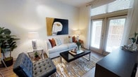 This single-floor space showcases a thoughtfully designed living area that balances contemporary style with a warm, inviting ambiance. The living room serves as the focal point, featuring smooth hardwood flooring that anchors the space with a rich, natural tone. A large glass-paneled door with white-slatted blinds floods the room with natural light, creating a bright and airy atmosphere while offering a visual connection to the outdoors.

The space is highlighted by clean white walls, which provide a crisp backdrop for a bold abstract artwork displayed prominently above the sofa—a striking combination of black, gold, and white tones that adds artistic character. The ceiling height contributes to an open, comfortable feel throughout the room.

Furnishings are carefully curated, including a cream-toned sofa accented with rust and patterned throw pillows, a sleek glass-and-metal coffee table, and a tufted charcoal accent chair. A gold-and-white patterned area rug defines the seating area and ties the design elements together cohesively. Matching chrome table lamps on either side of the room provide warm, balanced lighting, enhancing the polished, modern aesthetic of this well-appointed living space.