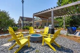Located at 900 Meadow Glen Pkwy in Fairburn, Georgia, this thoughtfully designed outdoor amenity space offers a welcoming retreat for community residents. At the heart of the area stands a substantial wood-beam pergola strung with globe lights, casting dappled shade over a expansive paver patio that serves as the central gathering hub. Beneath the pergola, a built-in stone outdoor kitchen with a grill provides an ideal setup for outdoor cooking and entertaining, while a plush blue sectional seating arrangement creates a comfortable lounge area with views of the surrounding greenery. Just beyond the pergola, a dedicated fire pit zone features a modern round fire bowl encircled by bright yellow Adirondack chairs set on a gravel pad, offering a cozy spot for relaxed conversation. A charming stone-accented gazebo structure adds architectural interest to the landscape nearby. The space is bordered by lush, mature trees, manicured pine straw beds, and open green lawns, providing a natural and serene backdrop. A fenced community pool is visible just beyond the pergola, seamlessly connecting the various outdoor amenities. Paved walkways tie the entire area together, making it easy to navigate throughout this inviting outdoor community space.