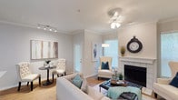 This single-floor residence in Lewisville offers a thoughtfully designed living space that balances comfort and style. The living area serves as the heart of the home, showcasing elegant crown molding, hardwood-style flooring, and smooth white ceilings accented by a multi-light ceiling fan. A white-tiled fireplace anchors one wall, adding warmth and a classic focal point to the room. Natural light pours in through multiple windows fitted with blinds, creating a bright and airy atmosphere throughout. A ceiling-mounted track lighting system provides additional illumination, further enhancing the open feel of the space. A cozy sitting nook near the entryway offers a charming secondary area, ideal for reading or casual conversation. The neutral color palette of soft grays and whites provides a clean, versatile backdrop that complements a wide range of décor styles. The overall ambiance is warm and inviting, with carefully considered architectural details such as the crown molding and fireplace surround lending a refined character to the space. This Lewisville residence combines functional layout with timeless design elements, making it a comfortable and visually appealing home.