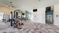 This single-level fitness center is thoughtfully designed to accommodate a full range of exercise needs within a spacious, open layout. The main workout floor features patterned tile flooring in a geometric design and is lined with an extensive selection of strength training equipment, including cable machines, resistance systems, free weights, and adjustable benches. Multiple wall-mounted televisions are positioned throughout for entertainment during workouts. A teal accent wall with warm sconce lighting and a built-in upholstered bench adds a distinctive design element, creating a welcoming focal point within the space. The cardio zone is outfitted with treadmills, ellipticals, rowing machines, and stationary bikes, all positioned in front of large windows that frame views of the surrounding outdoor pool area and community grounds, flooding the space with natural light. A dedicated cycling studio features vibrant green walls, light wood flooring, a ceiling fan, wall-mounted display, and a row of indoor cycling bikes — ideal for structured spin sessions. A teal tile water station with dual water fountains and a mirrored wall further enhance the facility's functionality. Direct access to the outdoor area is provided through glass double doors, seamlessly connecting interior and exterior amenities.
