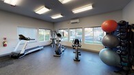 This single-floor fitness center at 9118 East Columbia Drive in Spokane, Washington offers a well-equipped workout facility designed for functional and comfortable exercise. The space features durable rubber flooring throughout, providing a safe and resilient surface for a variety of workouts. Large windows line the walls, flooding the room with natural light while offering views of the surrounding outdoor landscape, including green hillside scenery.

The cardio section is equipped with treadmills, an elliptical trainer, a stationary bike, and a rowing machine, giving users a full range of aerobic options. A multi-use cable and weight machine provides strength training capability alongside a dedicated area for functional fitness accessories, including a collection of exercise balls in multiple sizes. A wall-mounted flat-screen TV is positioned near the entrance for entertainment during workouts.

A full-length wall mirror enhances the sense of space and allows users to monitor their form during exercise. The room also includes a mini-split HVAC unit for climate control, bright overhead fluorescent lighting, and a Dasani water and beverage vending machine for post-workout refreshment. Coat hooks and a sanitizing station are conveniently located near the entry door, which features frosted glass etched with "Fitness Center." The overall ambiance is clean, bright, and purpose-built for an effective fitness experience.