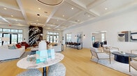 This expansive single-floor amenity space offers a sophisticated coastal-modern aesthetic across a generously sized open-concept layout. Light-toned hardwood flooring flows seamlessly throughout, unifying a series of distinct functional zones while maintaining an airy, cohesive atmosphere.

The main lounge area is anchored by a striking dark floor-to-ceiling fireplace surround, complemented by curated seating arrangements featuring leather and upholstered chairs beneath elegant circular pendant light fixtures. Coffered ceilings with recessed lighting add architectural depth and a refined finish overhead.

A dedicated co-working zone provides built-in desk stations set against a richly toned accent wall, offering a focused environment for productivity. Nearby, a casual dining and social area showcases a bold tiled accent wall, a marble-topped kitchen island, pendant lighting, and round dining tables with stylish seating.

Additional gathering areas include a bright sitting room with floor-to-ceiling black-framed glass doors and windows that flood the space with natural light and provide direct outdoor access. A game room section features a shuffleboard table beneath a shiplap ceiling, adding a relaxed recreational element.

Throughout, large windows and glass door entries ensure abundant natural light, while carefully selected artwork and décor contribute to a polished yet welcoming ambiance.