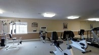 This single-floor fitness center offers a dedicated space for exercise and wellness. The room features warm beige walls accented by white baseboards, giving the space a clean and welcoming appearance. Flush-mounted square ceiling lights provide bright, even illumination throughout the area, ensuring excellent visibility for all workout activities.

The facility is equipped with a selection of cardiovascular and strength-training machines, including stationary bikes, a treadmill, and a multi-function cable weight machine, catering to a range of fitness routines. The light-colored speckled flooring is durable and easy to maintain, well-suited for a high-traffic exercise environment.

A "Fitness Rules" sign is prominently displayed on the wall, alongside a framed piece of artwork, adding a touch of structure and character to the space. A window along one wall allows natural light to supplement the overhead lighting, contributing to an open and energizing atmosphere. The layout is thoughtfully arranged to allow comfortable movement between equipment, making the space accessible and practical for users of varying fitness levels.
