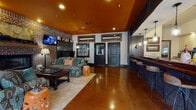 Located in Auburn, Alabama, this single-floor community space is designed with a warm, eclectic character that blends rustic charm with modern comfort. The main lounge area is anchored by a striking exposed brick wall featuring a wood-mantel fireplace, above which a wall-mounted television is perfectly positioned for communal viewing. Two richly upholstered sofas in bold ikat patterns face each other around a solid wood coffee table, creating an inviting seating arrangement. Warm amber-painted ceilings complement the polished concrete floors, which extend throughout the entire space and reflect the recessed and pendant lighting above.

Adjacent to the lounge is a generously sized bar area, outfitted with a deep navy base, a rich hardwood countertop, and a full row of industrial-style wood and metal barstools. Pendant lights with glass jar shades hang above the bar, illuminating the space with a warm glow. Behind the bar, a tile backsplash and open shelving provide a functional and visually appealing backdrop. A dedicated dining area along the windowed wall features multiple tables and chairs bathed in natural light through large windows dressed with plaid Roman shades, offering views of the surrounding exterior. The overall ambiance is lively, social, and thoughtfully appointed.