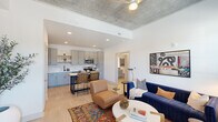 Located at 1045 Williamsburg Way in Evans, GA, this well-appointed one-bedroom, one-bathroom unit offers a thoughtfully designed open-concept layout across a single floor. The living room is a standout space, featuring a striking exposed concrete ceiling with a ceiling fan, light wood-style flooring, and large windows with natural light and outdoor views. The kitchen is modern and functional, outfitted with gray shaker-style cabinetry, gold hardware, quartz countertops, a hexagonal tile backsplash, stainless steel appliances, and a center island with bar seating — ideal for both cooking and casual dining. The living area flows seamlessly from the kitchen, creating an inviting, connected atmosphere. The bedroom offers a comfortable retreat with carpeted flooring, an exposed concrete ceiling detail, and windows with exterior views, complemented by a dedicated closet for storage. The bathroom features a clean, contemporary design with blue-gray cabinetry, a white quartz vanity, a lighted mirror, and a full tub-shower combination. A hallway connects the rooms efficiently, completing this compact yet well-organized layout that balances modern industrial design elements with warm, residential finishes throughout.