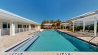Located at 3640 Blue Fin Way in Fort Myers, Florida, this vibrant outdoor amenity area delivers a true Florida resort lifestyle experience. At its centerpiece sits a generously sized swimming pool with crystal-clear blue water, flanked by a travertine paver pool deck complete with lounge chairs and a white pergola that provides welcome shade during sunny afternoons. Surrounding the pool, manicured tropical landscaping features tall swaying palm trees, well-trimmed hedgerows, and lush green lawn areas that create a private, resort-like atmosphere. A thatched tiki hut with pod-style lounge seating offers a relaxed retreat alongside the deck, while a dedicated sun-bathing platform lined with rows of white chaise lounges invites residents to soak up the Florida sunshine. A covered outdoor lounge area with comfortable seating and coastal-inspired décor provides a shaded gathering space. Stone paver pathways connect the various zones throughout the grounds, while cornhole boards and Adirondack chairs arranged on the lawn encourage casual outdoor recreation. The single-level clubhouse structure, finished in crisp white with a metal roof, anchors the space and opens directly to the amenity grounds, seamlessly blending indoor and outdoor living in classic Southwest Florida style.