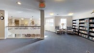 This inviting communal space is thoughtfully designed to encourage comfort and social interaction. The open-plan lounge area is anchored by plush seating arrangements, including classic plaid armchairs grouped around a side table and a cushioned sofa positioned beneath large landscape artwork, creating distinct yet connected areas for relaxation and conversation. Soft, wall-to-wall carpeting in a muted blue-grey tone runs throughout, contributing to the warm and quiet ambiance of the space.

A prominent white-railed balcony overlooks a lower-level dining area, where a statement chandelier hangs gracefully above a gathering of tables and chairs. Natural light filters in through multiple white-framed windows, brightening the neutral, cream-toned walls. Recessed and flush-mount ceiling lighting ensures the space remains well-lit throughout.

A well-stocked bookshelf lines one wall, offering a curated library corner complete with a floor lamp for reading. Decorative accents, including a console table with ornamental pieces and a traditional wall clock, add character and a homely feel. Structural columns are integrated seamlessly into the layout, and wide corridors extend from the lounge, providing easy accessibility and flow throughout the facility. Overall, the space exudes a calm, residential atmosphere suited to community living.