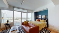 This thoughtfully designed single-floor residence combines modern aesthetics with functional living. The main living and sleeping area is a standout, anchored by expansive floor-to-ceiling sliding glass doors that flood the space with natural light and open to a balcony overlooking a broad cityscape. A bold teal accent wall frames the sleeping nook, complemented by warm-toned furnishings including a rich cognac leather sofa, a gold-hued bed, and a graphic black-and-white area rug that ties the space together. Track lighting on the crisp white ceiling adds a contemporary touch.

Adjacent to the main living area, a dedicated sitting nook features a wall-mounted television, a walnut-finished media console, and additional window views, providing a distinct space for relaxation. The kitchen is sleek and well-appointed, featuring two-tone cabinetry with dark walnut lower cabinets and light gray upper cabinets, white quartz countertops with under-cabinet LED lighting, a tile backsplash, stainless steel appliances, and warm hardwood flooring throughout.

The bathroom is clean and modern, finished with large-format neutral tiles, a glass-enclosed walk-in shower with full-height tile surround, a dark walnut vanity with a white countertop, an oversized mirror, and ample built-in storage. The overall ambiance is urban, stylish, and cohesive.