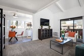 Located at 1942 Deerpark Dr in Fullerton, California, this single-story residence offers a well-organized layout designed for comfortable everyday living. The living room serves as the centerpiece of the home, showcasing vaulted ceilings with exposed beams that lend an airy, open quality to the space. Soft carpeting and crisp white walls create a clean, inviting atmosphere, while a large sliding glass door floods the room with natural light and provides direct access to a private balcony overlooking a landscaped communal setting.

The galley-style kitchen is efficiently designed with white cabinetry extending to the ceiling, granite-look countertops, wood-look flooring, and white appliances including a range and refrigerator. Recessed lighting ensures the space is well-illuminated throughout. Adjacent to the kitchen, the dining area features wood-look flooring, a ceiling fan, and a window that brings in additional natural light.

The bedrooms are comfortably sized with plush carpeting, ample natural light from large windows with vertical blinds, and easy access to closet space. The bathroom presents a clean, elongated layout with granite-look countertops, white cabinetry, a single sink, and wood-look flooring. Throughout the home, a cohesive neutral palette and consistent finishes contribute to a bright, well-maintained overall ambiance.