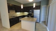 This kitchen is thoughtfully designed with a cohesive, modern aesthetic centered around rich, dark espresso cabinetry that extends to the upper and lower walls, providing generous storage throughout. The cabinetry is complemented by granite countertops featuring warm beige and gold tones, adding a polished, upscale feel to the space. A central island mirrors the granite surface and serves as a functional hub for meal preparation or casual dining, anchored by two elegant pendant lights that hang from the ceiling above.

The kitchen is equipped with a full suite of stainless steel appliances, including a refrigerator, range with electric cooktop, built-in microwave, and dishwasher, ensuring modern convenience. A undermount sink with a sleek faucet is positioned along the perimeter cabinetry. The backsplash features a neutral tile pattern that bridges the cabinetry and countertops cohesively.

Light tile flooring runs through the kitchen, transitioning to carpeting in the adjacent living area. Soft green-toned walls provide a subtle contrast to the darker cabinetry. A doorway visible in the background suggests access to additional rooms, rounding out this well-organized and visually appealing kitchen space.