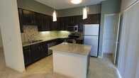 This kitchen is thoughtfully designed with a cohesive, modern aesthetic centered around rich, dark espresso cabinetry that extends to the upper and lower walls, providing generous storage throughout. The cabinetry is complemented by granite countertops featuring warm beige and gold tones, adding a polished, upscale feel to the space. A central island mirrors the granite surface and serves as a functional hub for meal preparation or casual dining, anchored by two elegant pendant lights that hang from the ceiling above.

The kitchen is equipped with a full suite of stainless steel appliances, including a refrigerator, range with electric cooktop, built-in microwave, and dishwasher, ensuring modern convenience. A undermount sink with a sleek faucet is positioned along the perimeter cabinetry. The backsplash features a neutral tile pattern that bridges the cabinetry and countertops cohesively.

Light tile flooring runs through the kitchen, transitioning to carpeting in the adjacent living area. Soft green-toned walls provide a subtle contrast to the darker cabinetry. A doorway visible in the background suggests access to additional rooms, rounding out this well-organized and visually appealing kitchen space.