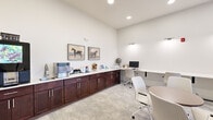 Located at 2755 Red Bloom Dr in Greenwood, Indiana, this single-floor office space is thoughtfully designed to support both focused work and casual collaboration. The interior is finished in a clean, neutral palette of crisp whites and light gray carpet, creating a bright and professional atmosphere. Recessed and wall-mounted lighting ensures the space is well-illuminated throughout.

Along one wall, rich dark-wood cabinetry with a white countertop serves as a dedicated beverage and resource station, complete with a coffee machine and ample counter space. Two framed equestrian art prints add a refined decorative touch above the cabinetry. Opposite, a long built-in white desk spans the wall beneath accent lighting, providing multiple workstations with ergonomic rolling chairs.

At the center of the room, a round table with seating offers a casual meeting area, while a pair of plush upholstered lounge chairs with warm orange accent pillows provide a comfortable conversation nook near the window. Natural light filters in through slatted blinds, offering views of the outdoors.

A floor-to-ceiling glass partition wall with a framed glass door visually opens the space to an adjacent area, enhancing transparency and connectivity. The overall design balances functionality with a welcoming, modern aesthetic.