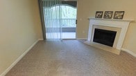 This single-floor living space offers a comfortable and functional layout centered around a well-appointed living room. The room features plush, neutral-toned wall-to-wall carpeting that adds warmth underfoot and complements the soft yellow walls throughout. A classic white-mantled fireplace with a dark interior surround serves as a focal point, adding both visual interest and a cozy ambiance to the space. Decorative wall art flanks the mantle, lending a personalized touch to the room.

Natural light flows generously into the space through a large sliding glass door dressed with vertical blinds, which opens directly onto a private balcony enclosed by a white railing. The balcony overlooks lush, mature greenery, providing a serene and private outdoor view that enhances the overall atmosphere of the interior. The arched window treatment above the sliding door adds an elegant architectural detail.

The overall ambiance is warm, neutral, and inviting, with the combination of natural light, soft carpeting, and the fireplace creating a relaxed and livable environment.