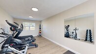 This dedicated fitness room offers a thoughtfully arranged workout space designed to support a well-rounded exercise routine. The room features light hardwood-style flooring throughout, with a gray exercise mat area designated for floor-based workouts and stretching. Neutral beige walls and flush circular ceiling lights create a clean, bright atmosphere that keeps the space feeling open and energetic.

Cardio equipment is a central focus, with two treadmills equipped with individual display screens positioned along one wall, alongside a stationary spin bike and an elliptical trainer. A large wall-mounted mirror on one side reflects the equipment and visually expands the room, making it easier to monitor form during workouts. A dedicated free weights section includes a tiered dumbbell rack stocked with hex dumbbells in multiple weights, paired with an adjustable workout bench set atop a protective mat. A vertical medicine ball rack lined with color-coded balls of varying weights adds versatility for functional training exercises.

Natural light enters through shuttered windows that offer views of the surrounding greenery, contributing to an inviting and motivating environment. A keyed entry door provides convenient direct access. The room is served by ceiling ventilation and is efficiently organized to maximize usable floor space without feeling cluttered, making it a practical and functional fitness facility.