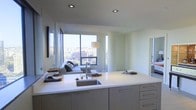 This contemporary single-floor residence offers a thoughtfully designed open-concept layout that seamlessly integrates the kitchen and living areas into one cohesive, flowing space. The kitchen is a standout feature, showcasing a large white waterfall-edge island with an undermount stainless steel sink, flat-panel white cabinetry, and recessed lighting overhead — all contributing to a clean, minimalist aesthetic. The kitchen opens directly into the living area, making the space feel generous and connected. Large, floor-to-ceiling windows flood the interior with abundant natural light and frame sweeping panoramic city views, giving the space an elevated, urban atmosphere. The neutral white palette throughout the walls, cabinetry, and countertops enhances the brightness and airiness of the interior. The living area is comfortable and inviting, with room for stylish furnishings that complement the modern design language. Recessed ceiling lights provide layered illumination throughout. The overall ambiance is polished and contemporary, ideal for those who appreciate clean lines, open spaces, and a strong visual connection to the cityscape beyond.
