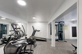 Located at Northtowne Village Apartments in Chattanooga, Tennessee, this dedicated fitness center offers a well-equipped workout space designed to meet a variety of exercise needs. The room is finished with clean white walls, neutral carpet tile flooring with teal accent borders, and flush circular ceiling lights that provide bright, even illumination throughout.

The cardio zone is anchored by Matrix-brand equipment, including a treadmill, elliptical, and stationary cycle, all positioned along a wall lined with large floor-to-ceiling mirrors that visually expand the space and allow users to monitor their form. A multi-station cable machine occupies the adjacent area, providing options for strength and resistance training, while a Matrix adjustable weight bench adds further versatility for free-weight workouts.

A wall-mounted flat-screen television is positioned at a comfortable viewing height, allowing residents to follow along with workout content or simply stay entertained during exercise sessions. The fitness center is accessed through a keyed entry door, and posted fitness room rules ensure a safe and organized environment for all users. The overall design is practical and modern, making efficient use of the available footprint to deliver a fully functional amenity for residents.
