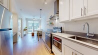 This well-appointed single-floor residence offers a thoughtfully designed open-concept layout that seamlessly connects the kitchen, dining, and living areas. The galley-style kitchen is outfitted with stainless steel appliances — including an electric range, dishwasher, and built-in microwave — complemented by white shaker cabinetry, open shelving, a stainless steel hood vent, and sleek countertops. Track lighting and a pendant light add functional and decorative illumination throughout the space.

The living area is bright and airy, bathed in natural light from large floor-to-ceiling sliding glass doors that open directly onto a private balcony, offering views of the surrounding urban streetscape. Light hardwood flooring runs consistently throughout the main living areas, adding warmth and cohesion to the interior.

The bedroom is a serene retreat, featuring a dedicated window with outdoor views, exposed ductwork as an industrial design accent, and ample space for furnishings. A hallway connects the bedroom to the full bathroom, which includes a white vanity with storage, large-format tile flooring, and a shower-tub combination. Conveniently located in the hallway is an in-unit stacked washer and dryer, along with additional storage shelving.