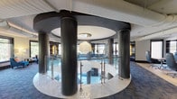 This architecturally distinctive single-floor space is defined by a striking circular atrium at its core, encircled by substantial dark cylindrical columns that rise to a recessed ceiling medallion. A dramatic cascading chandelier composed of layered glass or crystal elements serves as the focal centerpiece, suspended above a polished marble floor below. Sleek glass and stainless steel railings frame the atrium perimeter, lending a refined, contemporary aesthetic throughout.

Surrounding the central atrium, the space transitions into thoughtfully arranged lounge and workspace areas laid with deep blue patterned carpet. Multiple seating alcoves positioned along the perimeter benefit from expansive floor-to-ceiling windows, which draw in abundant natural light and offer views of surrounding greenery and urban streetscapes. Furnishings include plush velvet lounge chairs in jewel tones, neutral upholstered seating clusters, and individual desk workstations suited for professional use.

Dark wood and backlit built-in cabinetry elements punctuate the curved walls, providing both storage and visual depth. Exposed ductwork integrated into the ceiling adds an industrial counterpoint to the space's otherwise polished finishes. Signage visible on the atrium façade indicates a professional or mixed-use commercial setting. The overall ambiance is modern, upscale, and carefully curated.