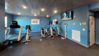 This fitness center offers a thoughtfully designed workout environment across a single level, combining functional layout with an inviting aesthetic. The space is defined by bold blue walls and a striking dark wood-plank ceiling with recessed lighting, creating a modern yet warm atmosphere throughout.

The main exercise area is generously sized, providing ample room for a comprehensive array of cardio equipment, including multiple treadmills, elliptical trainers, and stationary bikes. Wall-mounted flat-screen televisions are positioned for easy viewing during workouts. A dedicated strength and free weights section features a multi-station cable machine, a dumbbell rack, kettlebells, medicine balls, and a stability ball, catering to a wide range of training styles.

An adjacent room, accessible through glass-panel doors, features hardwood flooring and appears suited for group fitness classes or stretching. A water fountain is conveniently located near the entrance. Dark rubber flooring throughout the main workout area provides durability and safety underfoot. Framed athletic artwork adds visual interest to the walls, contributing to an energetic and motivating environment overall.