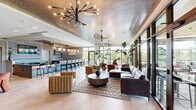 Located at 350 Carraway Crossing in Chapel Hill, NC, this thoughtfully designed amenity lounge offers a generous single-floor layout that seamlessly blends modern aesthetics with comfortable functionality. The space is anchored by a dramatic lounge area featuring an array of curated seating arrangements, including plush rust-colored velvet armchairs, a large leather sectional sofa, and a channeled caramel accent chair, all arranged around a round wood coffee table atop a textured area rug. Striking sputnik-style chandeliers and tiered pendant lighting fixtures illuminate the space from above, complemented by an intricately patterned textured ceiling that adds visual depth throughout.

Floor-to-ceiling glass curtain walls and expansive windows flood the interior with natural light and provide unobstructed views of the lush wooded landscape and serene pond beyond. A glass-railed outdoor balcony extends the living space outward, offering sweeping panoramic views of the surrounding North Carolina treeline. Large tropical plants positioned throughout enhance the biophilic design.

A dedicated bar and kitchen area serves as an entertainment hub, appointed with bold black marble countertops accented by white veining, vertical tile backsplash, open wood shelving, elegant teardrop pendant lights, a built-in wine refrigerator, and a draft beer tap. The light tile flooring ties the entire space together, creating a cohesive and inviting atmosphere throughout.