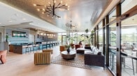 Located at 350 Carraway Crossing in Chapel Hill, NC, this thoughtfully designed amenity lounge offers a generous single-floor layout that seamlessly blends modern aesthetics with comfortable functionality. The space is anchored by a dramatic lounge area featuring an array of curated seating arrangements, including plush rust-colored velvet armchairs, a large leather sectional sofa, and a channeled caramel accent chair, all arranged around a round wood coffee table atop a textured area rug. Striking sputnik-style chandeliers and tiered pendant lighting fixtures illuminate the space from above, complemented by an intricately patterned textured ceiling that adds visual depth throughout.

Floor-to-ceiling glass curtain walls and expansive windows flood the interior with natural light and provide unobstructed views of the lush wooded landscape and serene pond beyond. A glass-railed outdoor balcony extends the living space outward, offering sweeping panoramic views of the surrounding North Carolina treeline. Large tropical plants positioned throughout enhance the biophilic design.

A dedicated bar and kitchen area serves as an entertainment hub, appointed with bold black marble countertops accented by white veining, vertical tile backsplash, open wood shelving, elegant teardrop pendant lights, a built-in wine refrigerator, and a draft beer tap. The light tile flooring ties the entire space together, creating a cohesive and inviting atmosphere throughout.