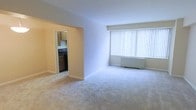 This single-floor residential space offers a straightforward and functional layout designed for comfortable everyday living. The main living area is finished with plush wall-to-wall carpeting in a neutral tone, creating a clean and understated foundation throughout. A large picture window dressed with vertical blinds dominates one wall of the living area, allowing ample natural light to fill the room while offering privacy control. Beneath the window, a baseboard heating unit ensures year-round comfort. The living space connects directly to a separate kitchen through an open doorway, providing an easy flow between the two areas. The kitchen features warm-toned wooden cabinetry and includes a dark-finish gas or electric range, providing essential cooking functionality. The overall color palette of the space is soft and neutral, with light-colored walls and trim throughout, contributing to a bright and airy ambiance. The single-floor plan makes the layout accessible and easy to navigate, with a cohesive design that prioritizes practicality. This space presents a solid foundation well-suited for personalization and customization to suit a variety of living styles.