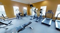 This fitness center is a dedicated, single-floor workout space designed to accommodate a range of exercise needs. The room features warm golden-yellow walls accented by crisp white crown molding and baseboards, creating an energizing yet welcoming atmosphere. Large windows on multiple walls flood the space with natural light, contributing to an open and motivating environment. The flooring is covered in durable gray gym-grade material, providing cushioning and traction suitable for active use. The facility is outfitted with a thoughtful selection of equipment, including a multi-function strength training machine, a weight bench, elliptical trainers, and a treadmill, offering users options for both cardiovascular and resistance workouts. A wall-mounted flat-screen television is positioned prominently, allowing exercisers to stay entertained during their sessions. A glass-paneled door provides access to an adjacent area, enhancing the sense of connectivity within the facility. The combination of functional equipment, ample natural light, and a clean, organized layout makes this fitness center a practical and inviting space for daily exercise.