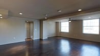 This single-floor space presents a clean, open layout designed with both comfort and functionality in mind. The main living area is anchored by rich, dark hardwood floors that run throughout, lending warmth and a sense of refined elegance to the interior. Neutral warm-toned walls complement the flooring and create a welcoming, versatile backdrop suitable for a variety of furnishing styles.

The ceiling features a combination of recessed lighting and a stylish ceiling fan, ensuring the space is well-lit and well-ventilated. Structural ceiling details, including a subtle overhead beam accent, add architectural interest and help define the open layout. A window along one wall invites natural light into the space, brightening the interior and adding an airy quality.

A doorway visible from the main area leads into a connecting hallway or adjoining room, hinting at additional spaces within the floor plan. White baseboards throughout provide a crisp contrast against the dark flooring, reinforcing the clean, finished aesthetic. Electrical outlets and HVAC vents are thoughtfully placed along the walls, reflecting attention to practical detail. Overall, the space offers a well-balanced blend of modern design and everyday functionality in a polished, move-in-ready presentation.