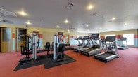 This fitness center offers a spacious, single-floor layout designed to accommodate a wide range of exercise needs. The interior features vibrant yellow and orange accent walls adorned with collegiate banners, giving the space an energetic and motivating atmosphere. The flooring combines red rubberized surfaces with black anti-fatigue mat sections beneath the strength training equipment, providing safety and comfort throughout.

The cardio zone is well-appointed with a row of treadmills equipped with individual digital display consoles, ideal for tracking workout metrics. Additional cardio machines are visible toward the rear of the space. The strength training area includes multi-function cable and weight machines, offering a variety of resistance training options for users of all fitness levels.

Overhead, ceiling fans aid in air circulation, complementing the recessed lighting and HVAC vents that maintain a comfortable environment. The textured ceiling and interior doors suggest a practical, community-focused design built for regular, high-traffic use. Overall, the fitness center is thoughtfully organized, balancing cardio and strength equipment within a bright, well-lit space that encourages an active and healthy lifestyle.