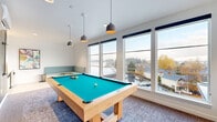 This single-floor game room is a well-appointed amenity space designed for recreation and socializing. The room is anchored by a solid wood-framed pool table with a vibrant teal felt surface, positioned centrally on a patterned area rug and illuminated by a trio of modern pendant lights. Large windows line the exterior walls, flooding the space with abundant natural light and offering expansive views of the surrounding waterway, marina, and neighboring rooftops.

A built-in banquette upholstered in a soft teal fabric runs along one wall, accompanied by small round pedestal side tables, creating a comfortable lounge area. Opposite the lounge, a casual dining nook features light wood-topped tables paired with olive green metal chairs, ideal for small gatherings. The flooring transitions between light hardwood planks and patterned carpet, adding texture and visual interest throughout.

A standout design feature is the dramatic floor-to-ceiling wood-grain accent wall, which serves as a backdrop for a wall-mounted pool cue rack. Adjacent to it, a compact kitchenette includes cabinetry, a sink, open shelving with patterned tile backsplash, and a mini refrigerator. Colorful wall art and navy roller shades complete the contemporary, inviting aesthetic of this versatile community space.