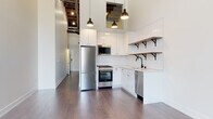 This single-floor loft-style space masterfully combines raw industrial character with contemporary design. The open living area commands attention with soaring ceilings that reveal original exposed wooden beams and structural elements overhead, while original brick walls add authentic texture and warmth. A large arched window with black-framed trim floods the space with natural light and offers views of the surrounding neighborhood, further enhancing the airy, expansive feel of the main living area. Dark-stained hardwood flooring runs throughout, grounding the space with a rich, cohesive look.

The kitchen is thoughtfully designed with white shaker-style cabinetry, a classic white subway tile backsplash, and open shelving with black iron brackets for both storage and display. Stainless steel appliances—including a French door refrigerator, gas range, built-in microwave, and dishwasher—provide a sleek, modern contrast to the kitchen's clean aesthetic. Industrial-style pendant lights hang above, tying into the loft's overall character.

The bathroom features a warm wood-toned vanity with a white countertop, black pull hardware, a tall matching storage column, a mirrored medicine cabinet, and classic white hexagonal tile flooring, delivering a refined yet timeless finish.