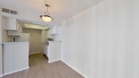 This single-story unit offers a functional and well-organized layout connecting a living area, kitchen, and bedroom. The living room is bright and open, featuring light wood-look flooring, a textured ceiling, and a flush-mount ceiling light. Vertical blinds on a sliding glass door allow natural light to filter in while providing privacy. The space flows directly toward the kitchen, which is separated by a half-wall peninsula with a laminate countertop—an efficient pass-through design that maintains openness between rooms.

The galley-style kitchen is efficiently laid out with white cabinetry lining both sides, complemented by light laminate countertops, a stainless steel sink, and white appliances including a refrigerator, electric range, and dishwasher. Overhead cabinetry provides ample storage. A door at the end of the kitchen leads to an additional utility or storage area.

The bedroom is spacious, finished with the same wood-look flooring, and features a ceiling fan with light fixture. A mirrored sliding closet door spans a large portion of one wall, reflecting light and adding depth to the room. Built-in shelving adjacent to the closet provides additional storage. A vanity area with white cabinetry is visible just off the bedroom, indicating a connected bathroom. Neutral wall tones and clean finishes give the entire unit a move-in ready feel.