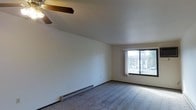 This single-floor space presents a clean, straightforward layout centered around a spacious living area. The room is finished with wall-to-wall carpeting in a neutral tone, providing a soft underfoot feel throughout. Overhead, a ceiling fan with a wooden beam accent offers both air circulation and a subtle decorative touch, keeping the space comfortable year-round.

Natural light enters generously through a large picture window fitted with sheer white curtains, which frame views of the outdoors, including visible greenery. The window contributes to an open, airy atmosphere while also allowing residents to adjust light levels as needed.

Along the base of one wall, a baseboard heater runs the length of the room, providing an efficient supplemental heat source during cooler months. The walls are finished in a clean, neutral white, offering a versatile backdrop suitable for a variety of interior styles.

Overall, the space maintains a simple, functional ambiance with practical features like the ceiling fan and baseboard heating integrated seamlessly into the design. The neutral palette and natural light make it an adaptable and welcoming environment.