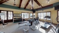 This fitness center is defined by its striking architectural design, most notably the soaring vaulted ceiling adorned with rich, dark-stained exposed wood beams that lend a lodge-like character to the space. Dual ceiling fans are mounted throughout, ensuring consistent air circulation. The upper walls are painted in a deep teal, transitioning to a warm cream below, creating a visually dynamic and inviting environment. Recessed lighting and large, wood-trimmed windows allow generous natural light to flood the interior, offering views of surrounding greenery.

The flooring consists of neutral, tile-patterned carpet throughout the main workout area. The space is comprehensively equipped with a strong mix of cardio machines, including Precor ellipticals, treadmills, and a stair climber. The strength-training zone features multiple cable and weight machines, a dedicated free-weight rack with dumbbells and medicine balls, a flat bench, and stability balls. Full-length mirrors are mounted along one wall, expanding the sense of space and supporting form monitoring. Entry is through elegant, dark wood-framed glass doors, reinforcing the refined aesthetic of this well-appointed facility.