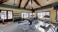 This fitness center is defined by its striking architectural design, most notably the soaring vaulted ceiling adorned with rich, dark-stained exposed wood beams that lend a lodge-like character to the space. Dual ceiling fans are mounted throughout, ensuring consistent air circulation. The upper walls are painted in a deep teal, transitioning to a warm cream below, creating a visually dynamic and inviting environment. Recessed lighting and large, wood-trimmed windows allow generous natural light to flood the interior, offering views of surrounding greenery.

The flooring consists of neutral, tile-patterned carpet throughout the main workout area. The space is comprehensively equipped with a strong mix of cardio machines, including Precor ellipticals, treadmills, and a stair climber. The strength-training zone features multiple cable and weight machines, a dedicated free-weight rack with dumbbells and medicine balls, a flat bench, and stability balls. Full-length mirrors are mounted along one wall, expanding the sense of space and supporting form monitoring. Entry is through elegant, dark wood-framed glass doors, reinforcing the refined aesthetic of this well-appointed facility.