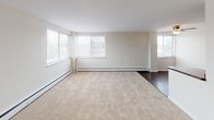 This single-floor residence offers a thoughtfully designed layout that balances comfort and functionality across its key living spaces. The main living area is generously sized, featuring wall-to-wall beige carpeting, neutral cream-toned walls, and multiple windows fitted with blinds that allow for abundant natural light. Baseboard heating runs along the perimeter, ensuring year-round comfort. The open feel of the living space is complemented by dark hardwood flooring that extends into the transition areas. The bedroom continues the warm, carpeted aesthetic and includes a ceiling fan with a light fixture, a window with a pleasant outdoor view, and its own baseboard heating unit, creating a cozy and private retreat. The kitchen is a functional galley-style space highlighted by warm honey-toned wood cabinetry, dark granite countertops, dark hardwood floors, and a full suite of black appliances including a refrigerator, range, microwave, and dishwasher. Track lighting overhead illuminates the workspace effectively. The bathroom is compact yet well-finished, featuring a dark espresso-toned vanity with a mirror cabinet, brushed nickel fixtures, and rich dark hardwood flooring, offering a clean and modern aesthetic. Overall, the space presents a neutral, move-in-ready environment with consistent finishes throughout.