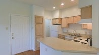 This single-story space showcases a well-designed kitchen as its focal point. The kitchen features light natural wood cabinetry that lines the upper and lower walls, providing ample storage. White appliances, including a refrigerator, dishwasher, and stovetop, are seamlessly integrated into the layout. A curved peninsula with a light-toned countertop extends into the living area, offering additional workspace and casual seating potential. Recessed lighting in the ceiling provides consistent, even illumination throughout the kitchen area. A stainless steel sink is positioned along the perimeter countertop, accompanied by a standard faucet. The flooring throughout is a warm hardwood-style material that adds a natural, cohesive aesthetic to the space. Neutral white walls keep the interior feeling bright and open. A paneled door with a distinctive two-panel design is visible, adding a subtle architectural detail. The layout is practical and efficient, with clear sightlines from the kitchen into adjacent areas, contributing to an open, connected feel throughout the single-floor residence.