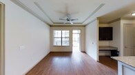This single-story, two-bedroom, two-bathroom residence offers a thoughtfully designed layout with refined finishes throughout. The living area serves as the heart of the home, showcasing rich hardwood-style flooring that extends seamlessly across the space. A decorative tray ceiling with crown molding adds architectural depth and elegance, while a ceiling fan provides both style and functional comfort. Natural light flows in through a large window fitted with blinds, and a glass door leads directly to a private balcony, offering an easy indoor-outdoor connection. The neutral, light-toned walls create a clean and versatile backdrop suited to a variety of décor styles. Adjacent to the living area, the kitchen is visible and features dark cabinetry and stone-look countertops, providing a striking contrast to the lighter tones of the main living space. The columned transition between rooms adds a subtle architectural detail that enhances the overall sense of structure and design. With two bedrooms and two full bathrooms, this residence provides ample space for comfortable everyday living.