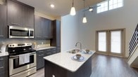 This two-story residence showcases a thoughtfully designed interior with modern finishes throughout. The kitchen serves as a central highlight, featuring floor-to-ceiling dark wood-grain cabinetry, speckled granite countertops, and a full suite of stainless steel appliances including a range, over-the-range microwave, and refrigerator. A kitchen island with an integrated sink provides additional workspace and casual dining potential, illuminated by stylish pendant lighting overhead. A tiled backsplash adds a refined detail to the cooking area. Wood-look flooring flows through the main living level, where an open staircase with a sleek metal railing connects to the upper floor.

The bedroom is notably spacious, benefiting from impressively tall ceilings that create an airy atmosphere. Large French-style doors and a clerestory window allow generous natural light to fill the room. A walk-in closet with built-in shelving and multiple hanging rods provides ample storage. The bathrooms are finished with granite-topped dark vanities, large framed mirrors with well-placed vanity lighting, and full tub-shower combinations, maintaining the home's consistent contemporary aesthetic throughout.