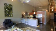 This single-floor space showcases an open-concept layout that seamlessly connects the living and kitchen areas, creating a bright and airy atmosphere. The kitchen is a standout feature, highlighted by rich warm-toned wood cabinetry that extends to impressive heights, complementing the vaulted ceilings above. A peninsula with granite countertops serves as a natural dividing point between the kitchen and living space, offering bar-height seating with modern stools — ideal for casual dining or entertaining.

Three elegant pendant lights hang above the peninsula, providing focused task lighting, while multiple ceiling fans with integrated lighting ensure comfort throughout the space. Stainless steel appliances, including a full-size refrigerator, electric range, and microwave, add a contemporary, professional-grade feel to the kitchen.

The polished concrete floors run throughout, contributing to the modern industrial character of the space. Neutral gray walls keep the palette versatile and clean. The adjacent living area offers a relaxed, comfortable setting with a glass coffee table and ample seating. The high, angled ceilings amplify the sense of spaciousness throughout this well-designed, cohesive single-level layout.