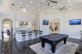 Located at 29980 FM-2978 in Magnolia, Texas, this sophisticated single-story facility presents an impressive blend of classical architectural design and modern amenities. The grand entrance lobby sets the tone with its soaring ceilings, large-format tile flooring, and a striking glass-top centerpiece table atop an ornate area rug, flanked by an adjacent reception desk. Arched doorways and fanlight windows are recurring architectural details throughout, lending the space a refined, stately character.

The expansive lounge area features rich, dark hardwood floors, coffered ceilings with recessed lighting, and a wall of tall arched windows that flood the room with natural light and offer views of the surrounding landscape. A fireplace anchors one end of the lounge, complemented by a beverage station and wall-mounted display screen, while generous seating arrangements invite gathering and relaxation.

The gourmet kitchen is outfitted with dark granite countertops, white cabinetry, a professional-grade gas range with a stainless steel hood, and a spacious island with bar seating. A dedicated games room provides additional entertainment, housing a full-size billiards table, multiple wall-mounted screens, and ample natural light from high clerestory windows. Throughout, the design balances grandeur with warmth, creating a welcoming and functional environment.