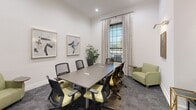 Located at 14086 Airline Hwy in Gonzales, Louisiana, this polished meeting room offers a well-appointed setting designed for productive gatherings and professional discussions. The space is anchored by a substantial rectangular conference table surrounded by ergonomic mesh-back chairs with chartreuse green upholstered seats, providing comfortable seating for multiple attendees. A sage green lounge chair in the corner adds a relaxed secondary seating option.

The room features crisp white walls adorned with framed abstract artwork, complemented by crown molding that lends a refined architectural touch. A large multi-pane window dressed with elegant linen drapes floods the room with natural light and offers views of the surrounding outdoor area, creating an open and airy atmosphere.

Built-in storage and display elements enhance the room's functionality and aesthetic appeal. A tall dark cabinetry unit with open shelving on one wall provides organized storage, while a recessed niche with warm-lit wood shelving displays curated decorative accents and a charcoal lower cabinet beneath. The room is accessed through a classic French door with glass panes, adding sophistication and visual connectivity to the adjoining space. Plush patterned carpet underfoot completes the professional yet inviting ambiance of this thoughtfully designed room.