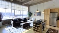 This single-floor loft showcases a striking industrial aesthetic defined by exposed concrete ceilings and structural elements that lend the space a raw, urban character. The open-concept layout seamlessly blends the living and dining areas, creating a generous and functional flow throughout. Floor-to-ceiling grid-pane windows dominate one wall, flooding the interior with natural light and offering open views of the surrounding cityscape. The living area is anchored by a large sectional sofa and complemented by a white accent chair, both arranged over a bold chevron-patterned area rug. Decorative framed art posters add a touch of personality to the walls. Adjacent to the living space, a bar-height table with a sleek dark countertop serves as a dining and entertaining hub, complete with pendant lighting overhead and a built-in wine rack below for storing bottles with ease. Bar stools tuck neatly underneath, making it ideal for casual dining or hosting guests. Lush indoor plants enhance the space with organic warmth, softening the contrast of the industrial concrete surroundings. The overall ambiance is modern, comfortable, and thoughtfully curated, balancing utilitarian design with welcoming, stylish furnishings.