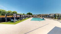 Located at 100 Hive Dr in Charlotte, NC, this single-level outdoor amenity complex is designed to deliver a resort-style experience within a residential community. At its centerpiece is a generously sized swimming pool with clear, turquoise water, surrounded by a wide concrete deck finished with light-toned pavers. Lounge chairs and market umbrellas are arranged along the poolside, offering ample space for sunbathing and relaxation under open Carolina skies.

Adjacent to the pool, a row of stylish private cabanas lines a lush synthetic turf lawn. Each cabana features a dark pergola frame with draped curtains, lounge seating, and side tables, providing shaded retreats throughout the day. A covered outdoor seating nook on the side of the main building showcases a bold, colorful mural, adding an artistic focal point to the landscaped grounds, which are accented with evergreen shrubs and decorative rock beds.

The support building is finished in crisp white board-and-batten siding and houses restroom facilities and water fountains, conveniently accessible from the pool deck. A dedicated pickleball court, enclosed by chain-link fencing, rounds out the active recreation offerings. Well-maintained walkways and ample parking complete this thoughtfully designed community amenity space.
