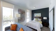 This single-floor space showcases a thoughtfully designed bedroom that balances modern aesthetics with comfortable functionality. The room's most striking feature is a deep charcoal accent wall, which serves as a dramatic backdrop for a colorful, abstract piece of artwork, adding a vivid focal point to the space. Opposite the accent wall, large windows fitted with roller shades allow generous natural light to fill the room, contributing to an open and airy feel.

The bedroom comfortably accommodates a full-sized bed with a dark wood headboard, dressed in neutral, layered bedding accented with pops of yellow and green. A light grey upholstered sofa positioned near the windows introduces a seating area, adorned with orange throw pillows that echo the warm tones found in the wall art. The flooring features wood-look planks that add warmth and texture underfoot.

A contemporary floor lamp with a classic drum shade provides supplemental lighting, while a geometric wire wall sculpture adds an artistic, three-dimensional element to the white walls. The overall ambiance is modern and curated, with a cohesive use of contrasting tones and carefully selected décor creating a stylish, livable environment.