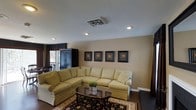 Located at 700 N Spence Avenue in Goldsboro, North Carolina, this single-story property offers a comfortable and well-appointed living environment. The main living area is designed with a warm, neutral color palette, featuring beige walls that create an inviting and cohesive atmosphere. Recessed lighting is installed throughout the ceiling, providing even, ambient illumination complemented by a central ventilation unit integrated seamlessly into the ceiling design. The living room is spacious enough to accommodate a large sectional sofa arrangement, centered around a classic dark wood coffee table and anchored by a traditional patterned area rug. A series of four framed botanical artworks lines the main wall, adding a decorative and coordinated visual element. A tall, dark armoire stands in the corner near a window dressed with structured blinds and draping curtains, offering both storage and style. Dark hardwood flooring runs throughout, providing a rich contrast to the lighter furnishings. Natural light filters in through the window, adding brightness to the space. The overall ambiance is warm, classic, and functional, making this property a well-rounded and comfortable single-floor home in Goldsboro.